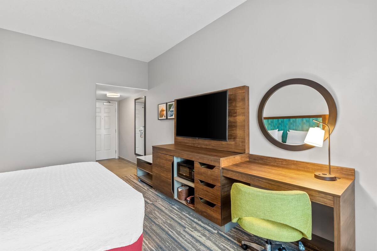 A hotel room with a bed, a desk with a TV, a mirror, and a chair.