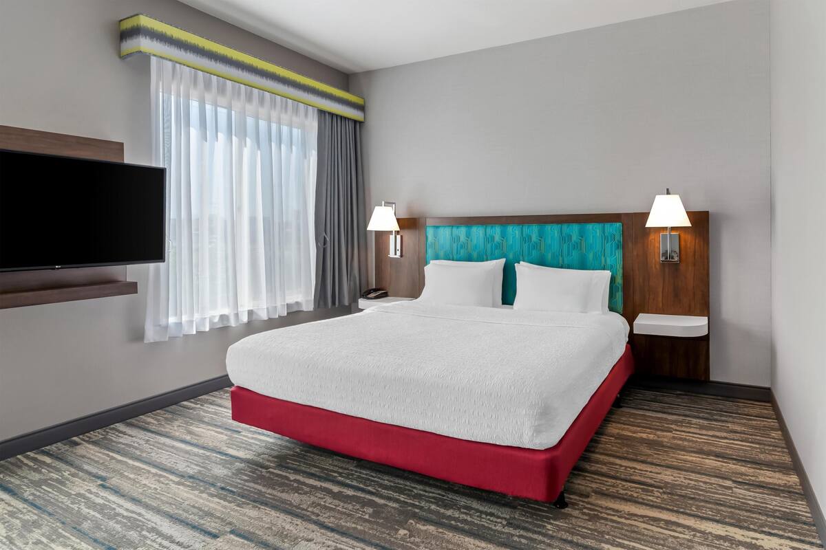 A hotel room with a large bed, two bedside lamps, a flat-screen TV mounted on the wall, and a window with sheer curtains.