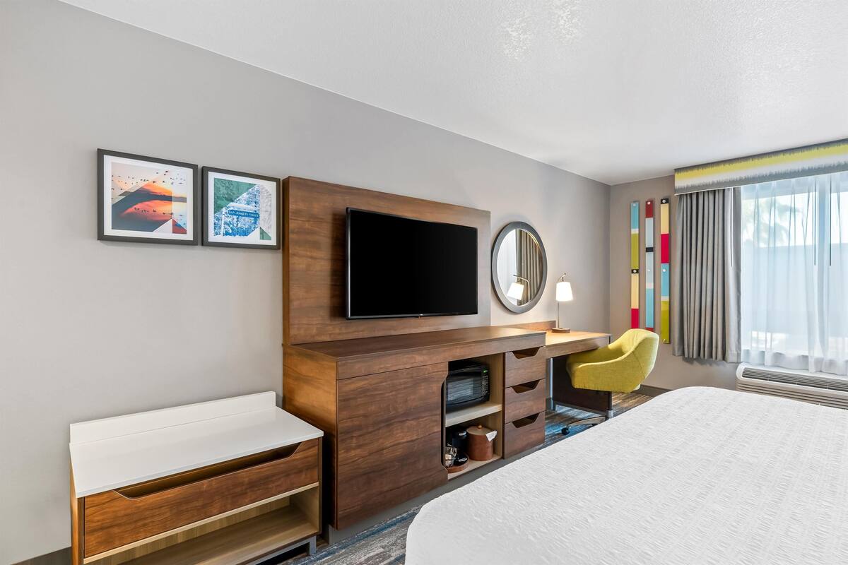 A hotel room with a bed, a desk with a chair, a flat-screen TV, a mirror, and two framed pictures on the wall.