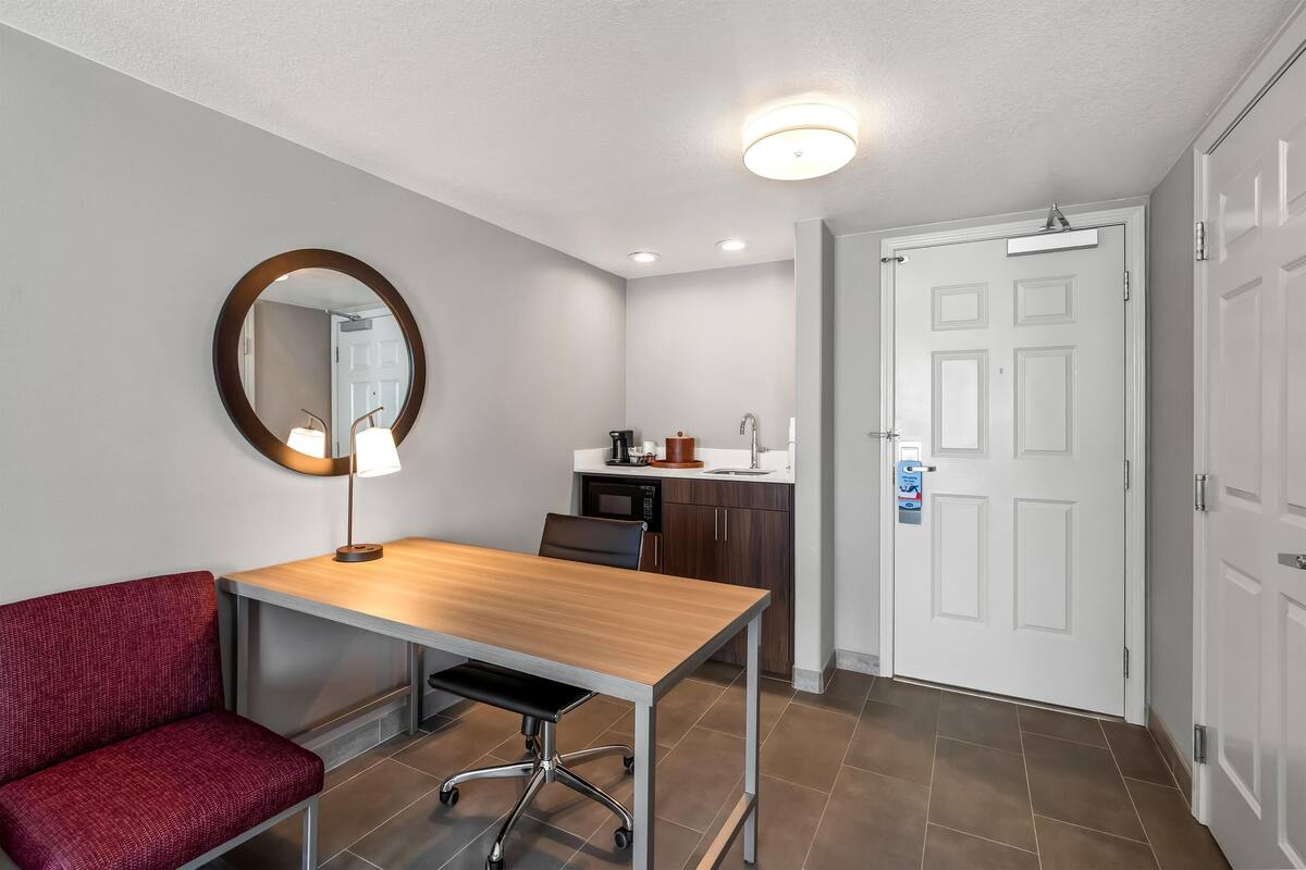 A hotel room with a wooden desk, a red upholstered bench, a round mirror, a white door, and a kitchenette area.
