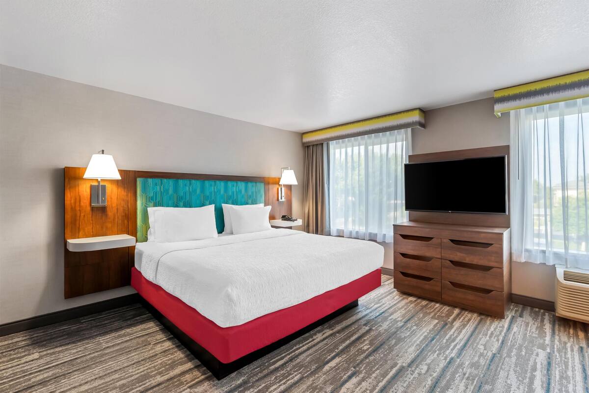 A hotel room with a large bed, a flat-screen TV, a wooden dresser, and a window with curtains.