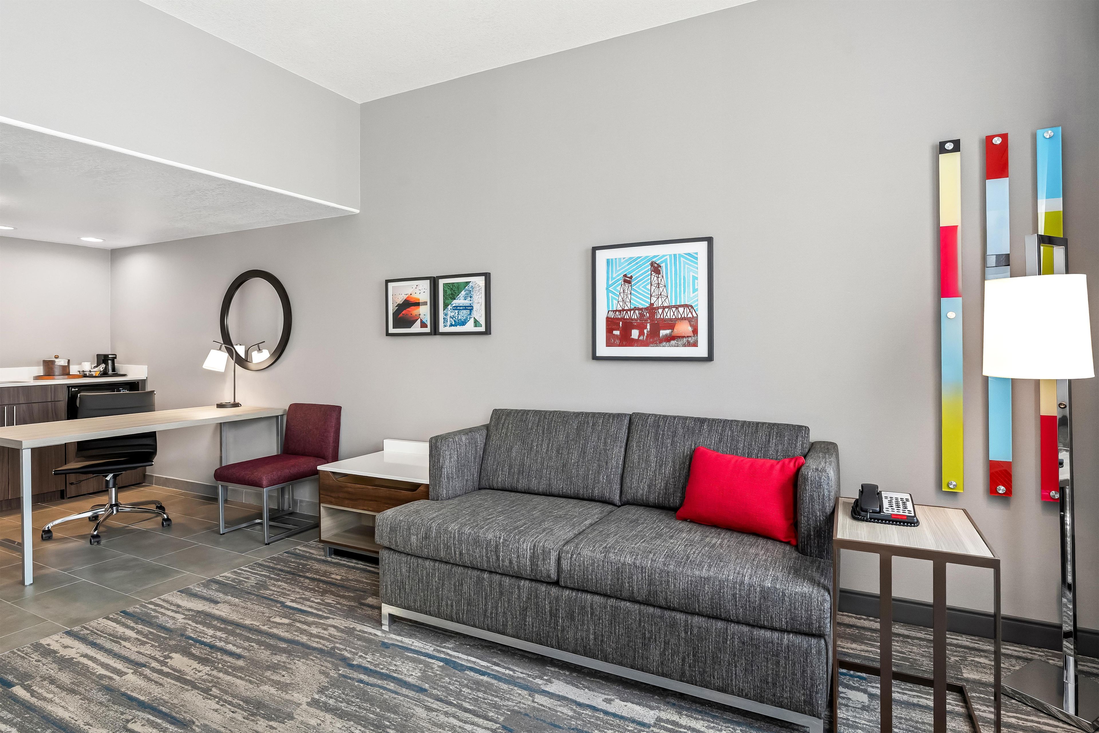 A modern living room with a grey sofa, red cushion, and a coffee table. There are two framed artworks on the wall, a desk with a chair, and a lamp on a side table.