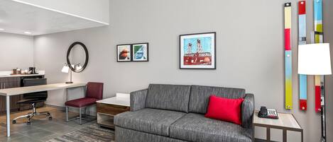 A modern living room with a grey sofa, red cushion, and a coffee table. There are two framed artworks on the wall, a desk with a chair, and a lamp on a side table.