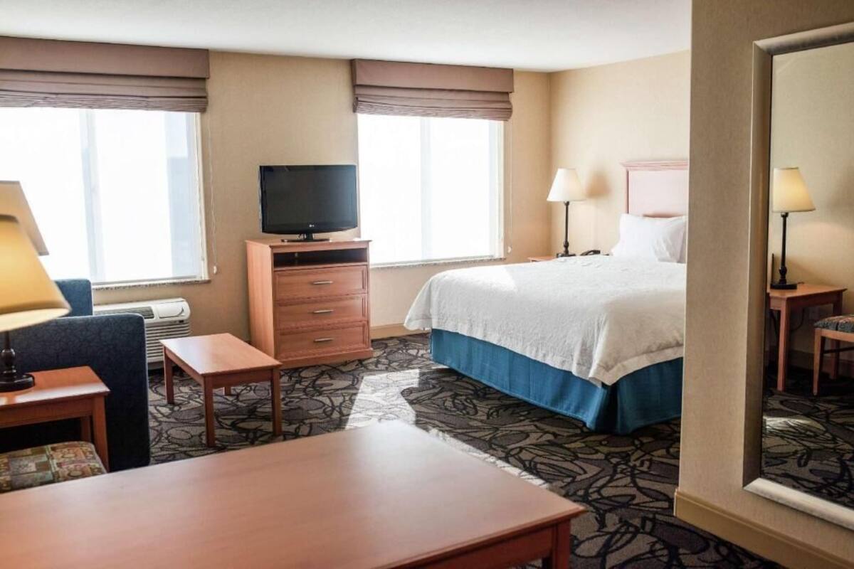 A hotel room with a bed, a television on a stand, a desk, a chair, and a window with curtains.