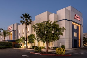 Un hotel Hampton Inn & Suites con fachada moderna, palmeras y un letrero que dice 'No Parking Fire Lane'.