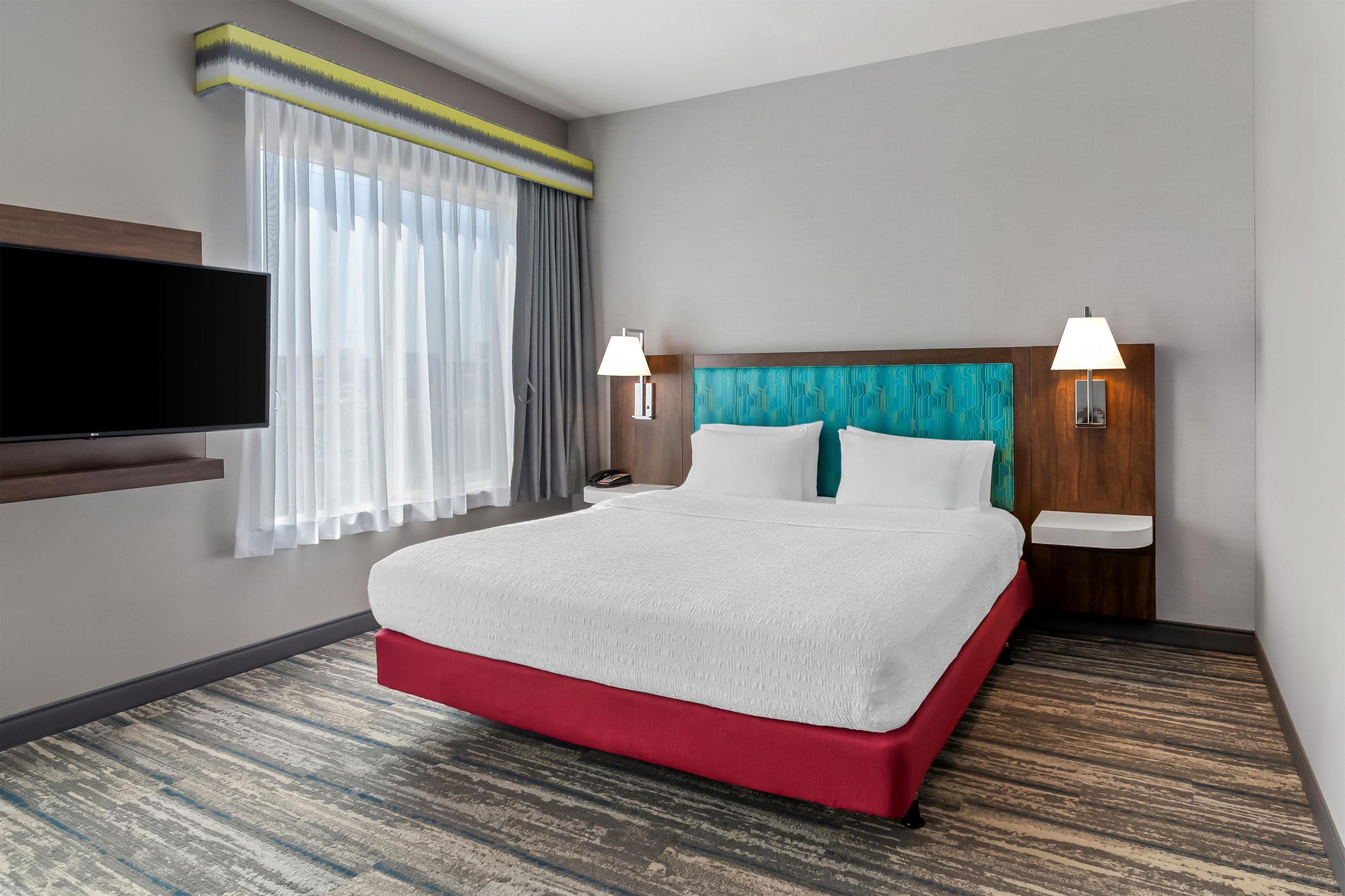 A hotel room with a bed, a flat-screen TV, a nightstand, a lamp, and a window with curtains.