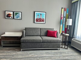 Una habitación de hotel moderna con un sofá gris, un escritorio pequeño y una lámpara colorida. Hay tres obras de arte enmarcadas en la pared.