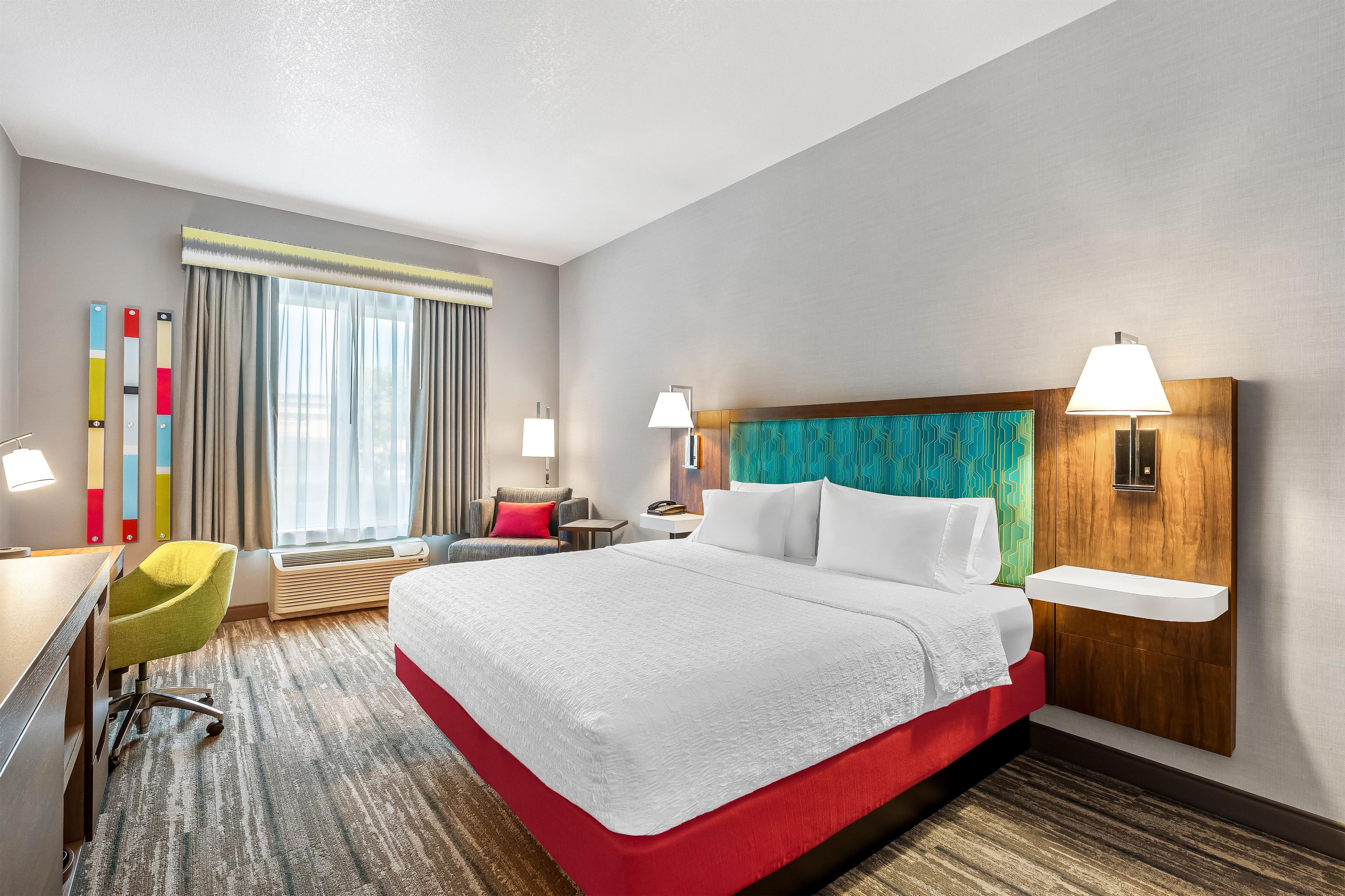 A hotel room with a large bed, a desk, a chair, a desk lamp, a bedside table, and a window with curtains.