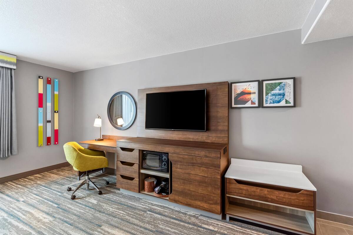 A modern hotel room with a wooden desk, a flat-screen TV, a yellow chair, a round mirror, and two framed artworks on the wall.