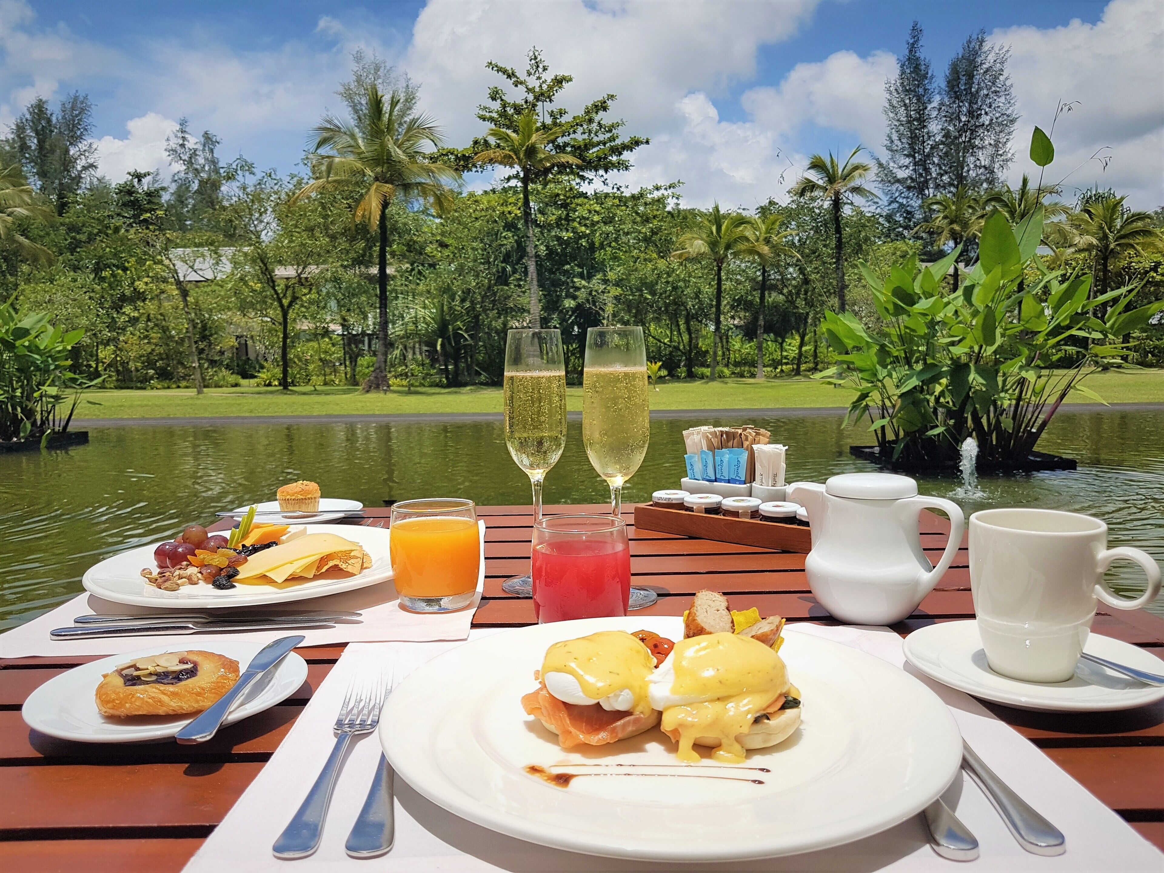 Un desayuno con una mesa junto a un lago, que incluye huevos Benedict, tostadas, fruta y jugo.