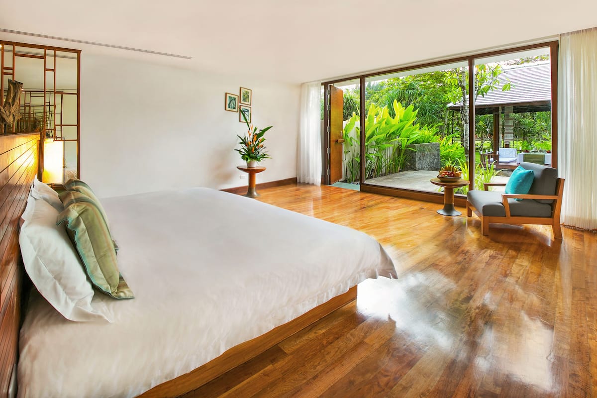 Un dormitorio con una cama grande, suelo de madera, puerta corredera de vidrio que da a un jardín y una zona de estar con una mesa y sillas.