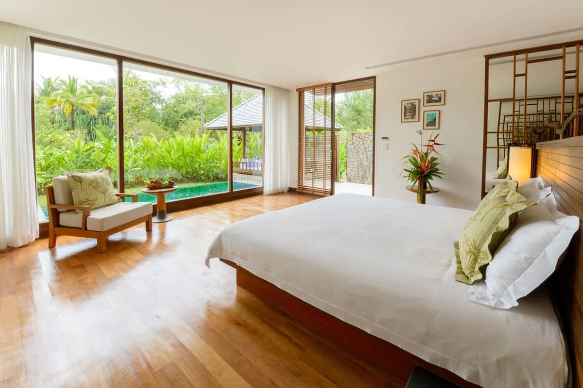 Un dormitorio amplio con una cama grande, suelo de madera, una zona de estar con un banco y vistas a vegetación exuberante a través de amplios ventanales.