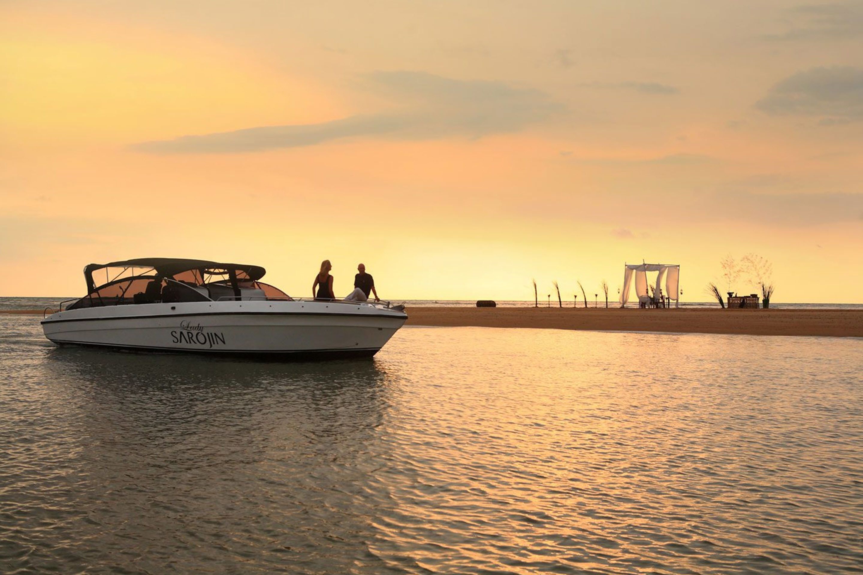 A boat named 'Lady SAROJIN' on calm water with a beach and structures in the background.