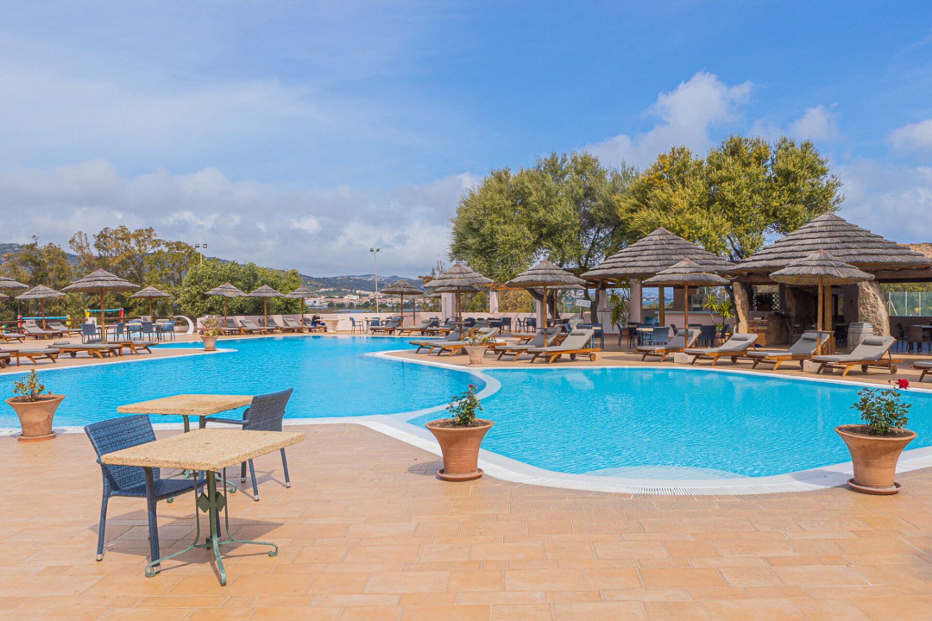A swimming pool with lounge chairs and umbrellas, surrounded by potted plants and a clear sky.