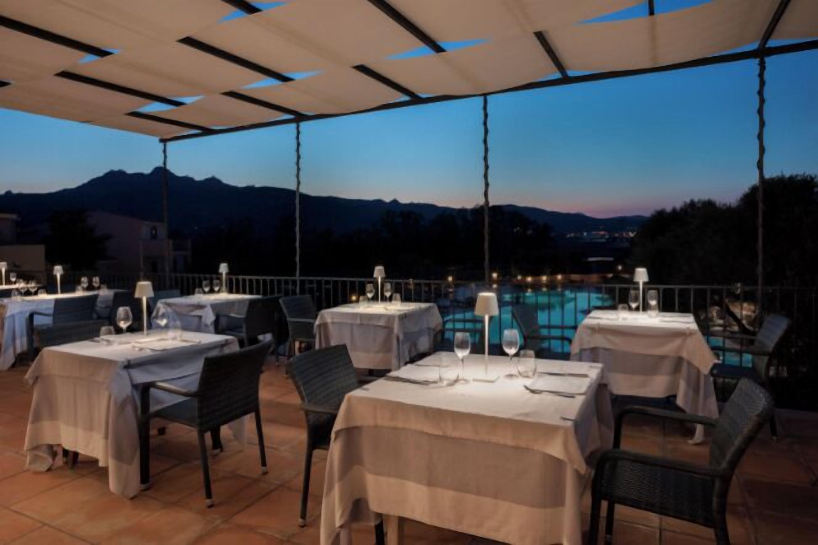 An outdoor dining area with tables and chairs, a pool in the background, and mountains in the distance.