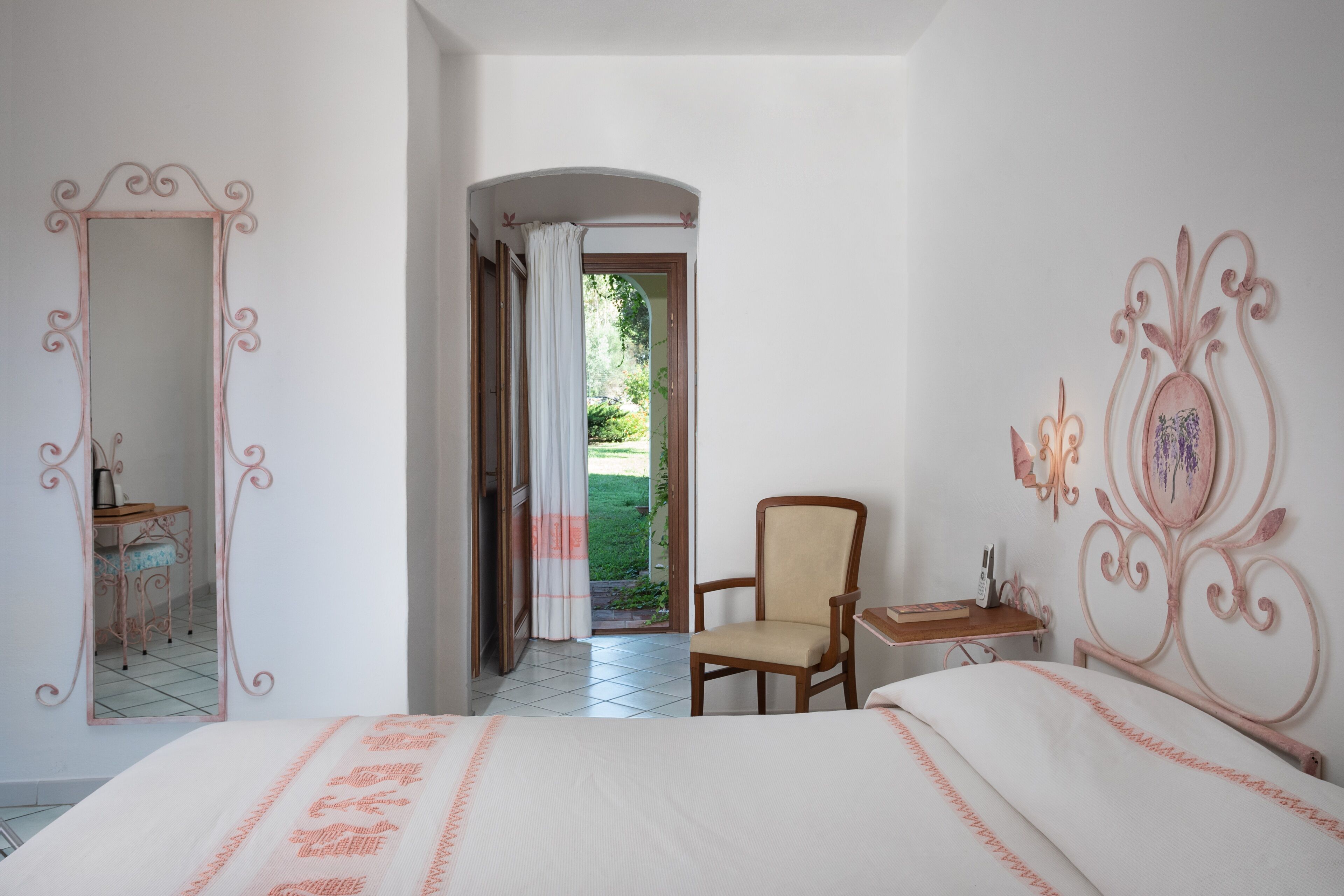 A hotel room with a bed, a chair, a vanity with a mirror, and a door leading to a garden.