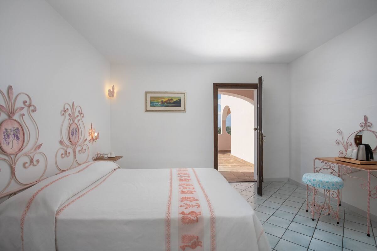 A bedroom with a bed, a desk, a chair, a picture on the wall, and a door leading to an outdoor area.