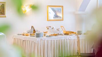 Mesa de buffet com diversos alimentos e um laptop, em um salão com paredes brancas e quadros nas paredes.