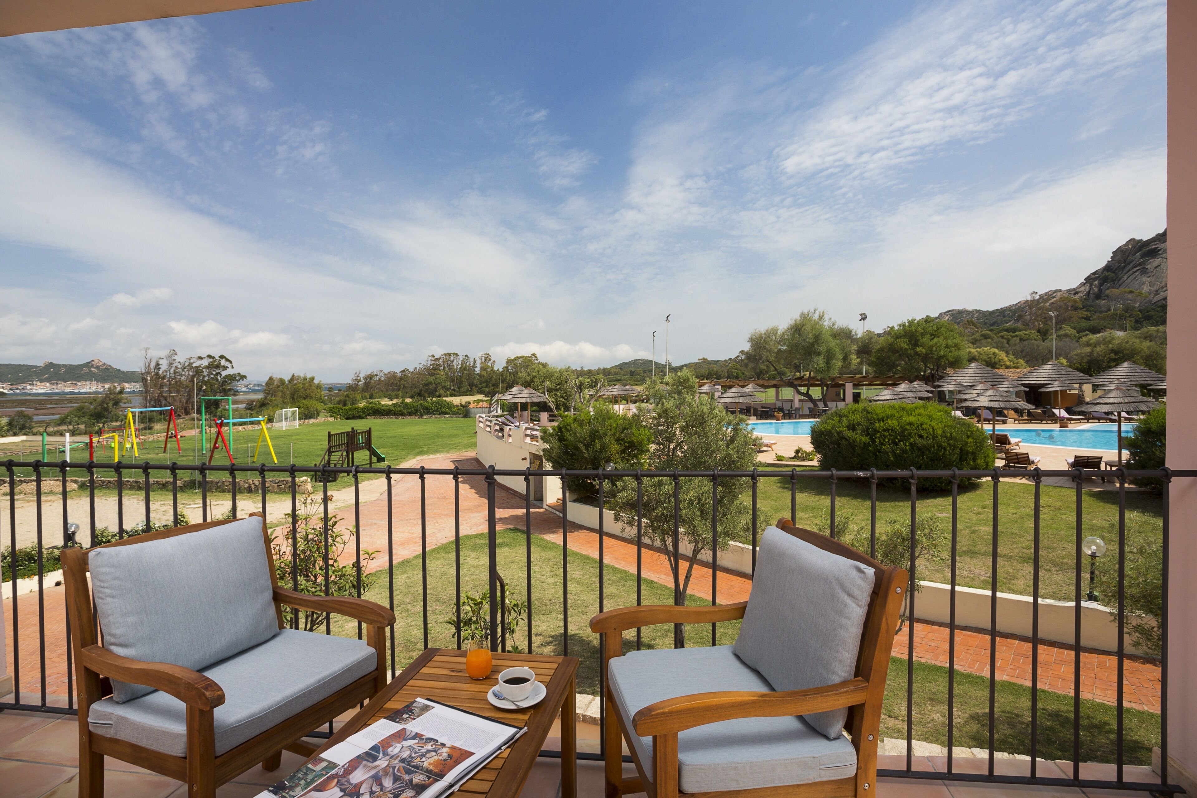 Un balcone con due sedie di legno e un tavolo, con vista su una piscina e un parco.