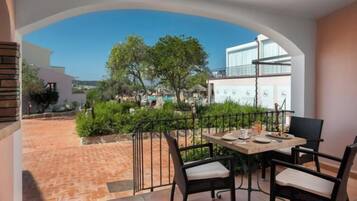 Een balkon met een tafel en stoelen, uitzicht op een zwembad en groen, en een gebouw op de achtergrond.