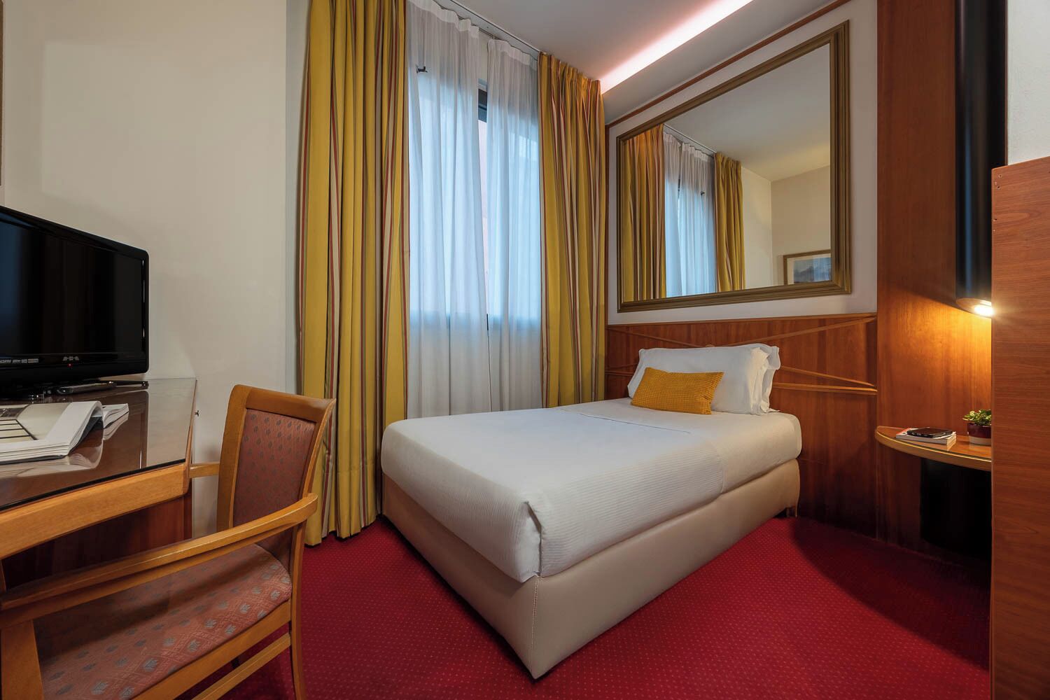Habitación de hotel con cama, escritorio con televisor, una silla, espejo y alfombra roja.