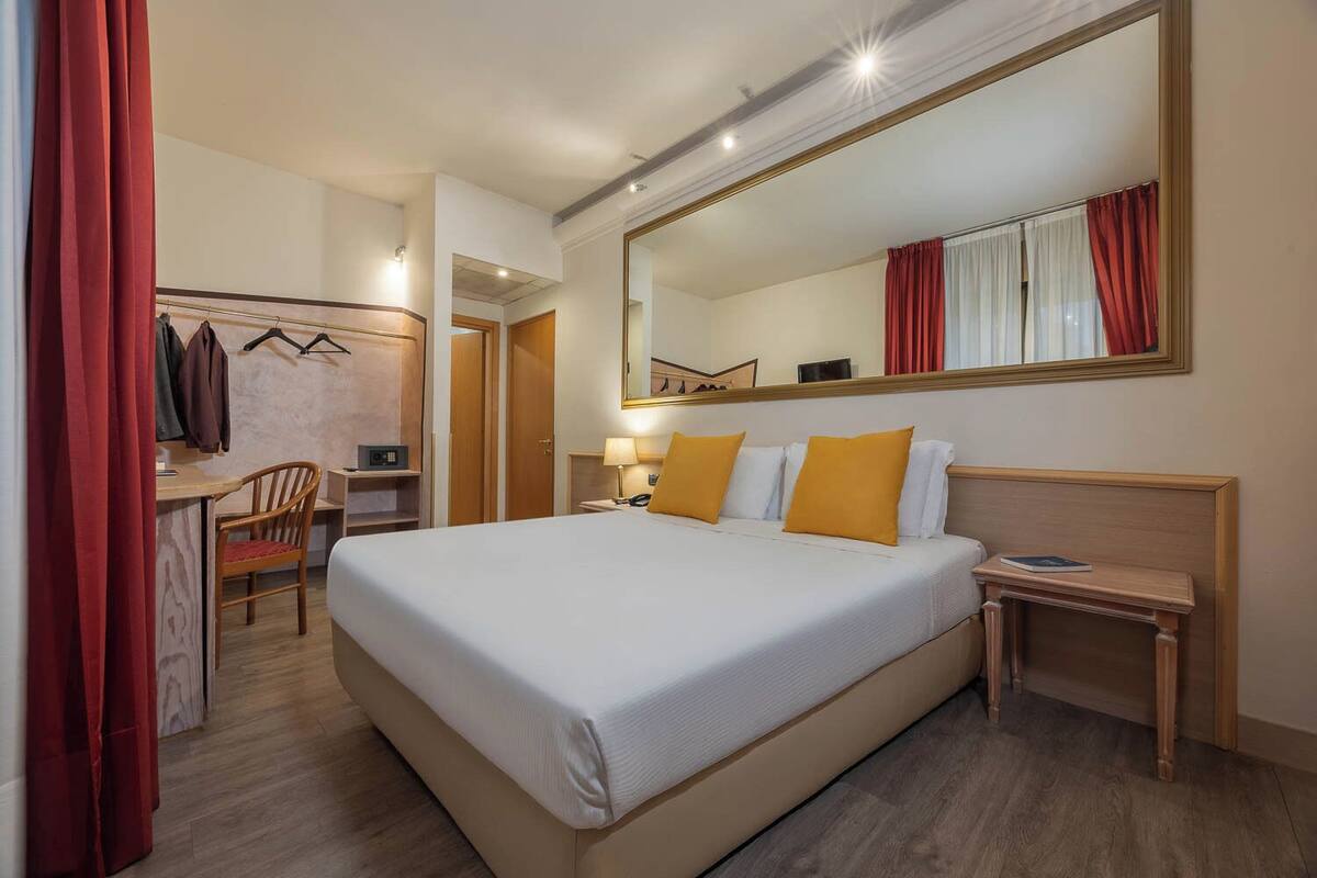 Ein Hotelzimmer mit einem großen Bett, einem Schreibtisch mit Stuhl, einem Spiegel und einem Nachttisch mit Lampe.