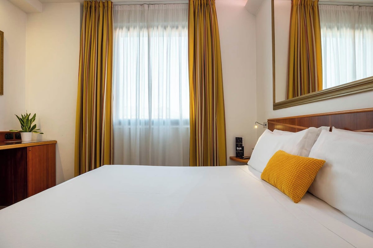 Une chambre d’hôtel avec un grand lit, une table de chevet avec une lampe, une petite plante en pot et un miroir au mur.