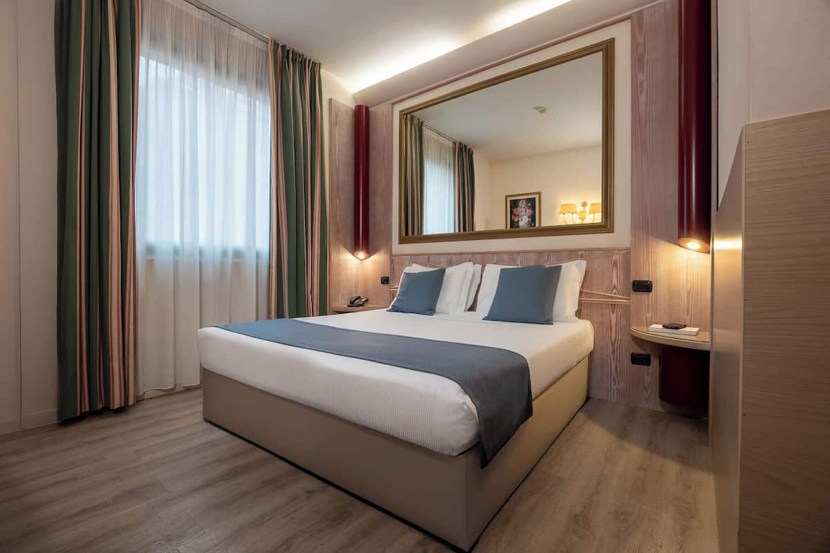 Ein Hotelzimmer mit einem großen Bett, einem Nachttisch mit Lampe, einem Spiegel und einem Holzboden.