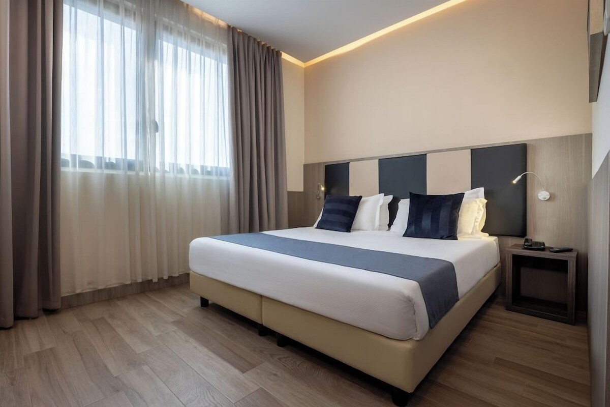 A hotel room with a large bed, a bedside table, a nightstand, and a window with curtains.