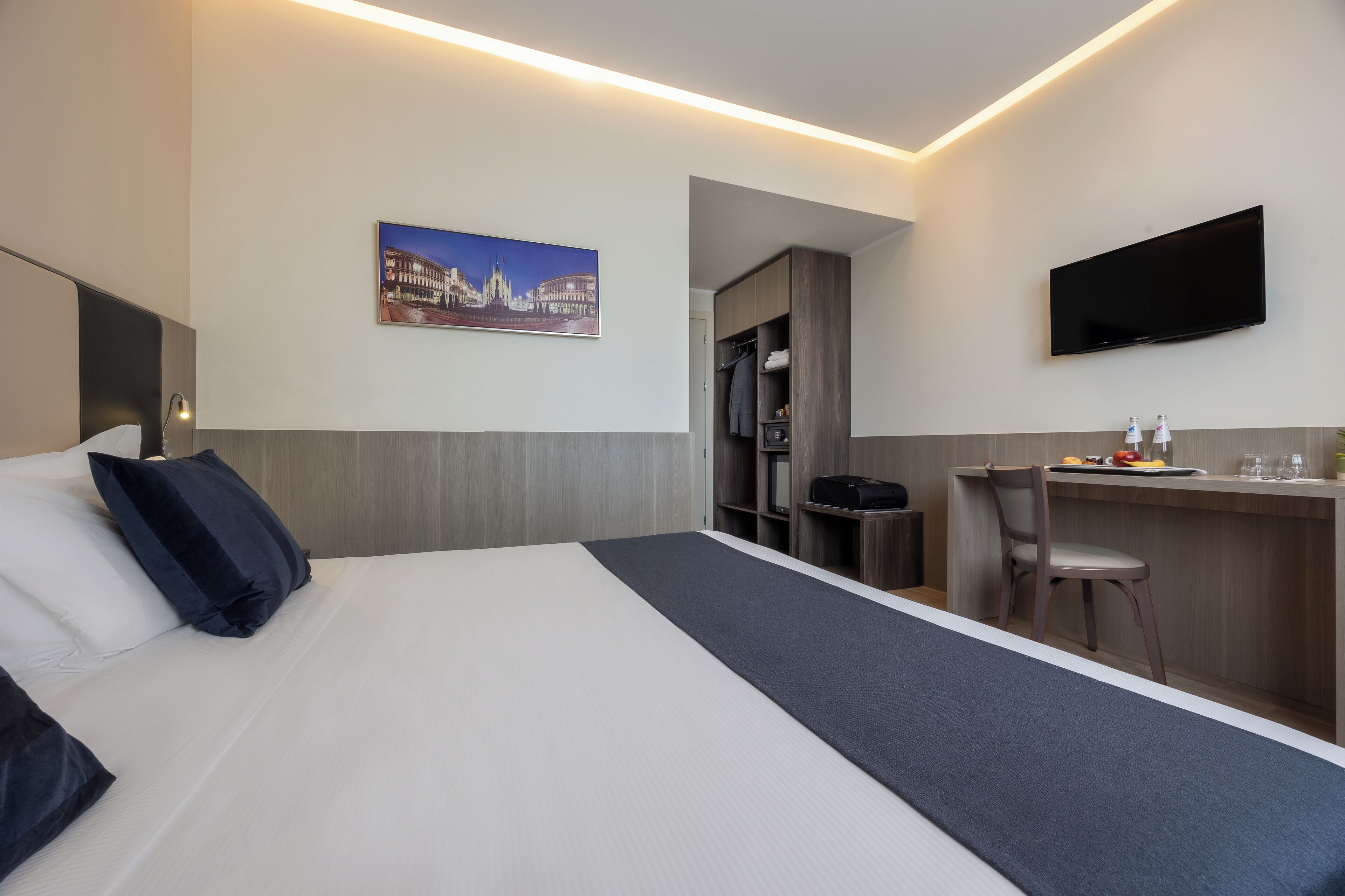Hotellihuone, jossa on sänky, työpöytä, tuoli, televisio ja taulu seinällä.