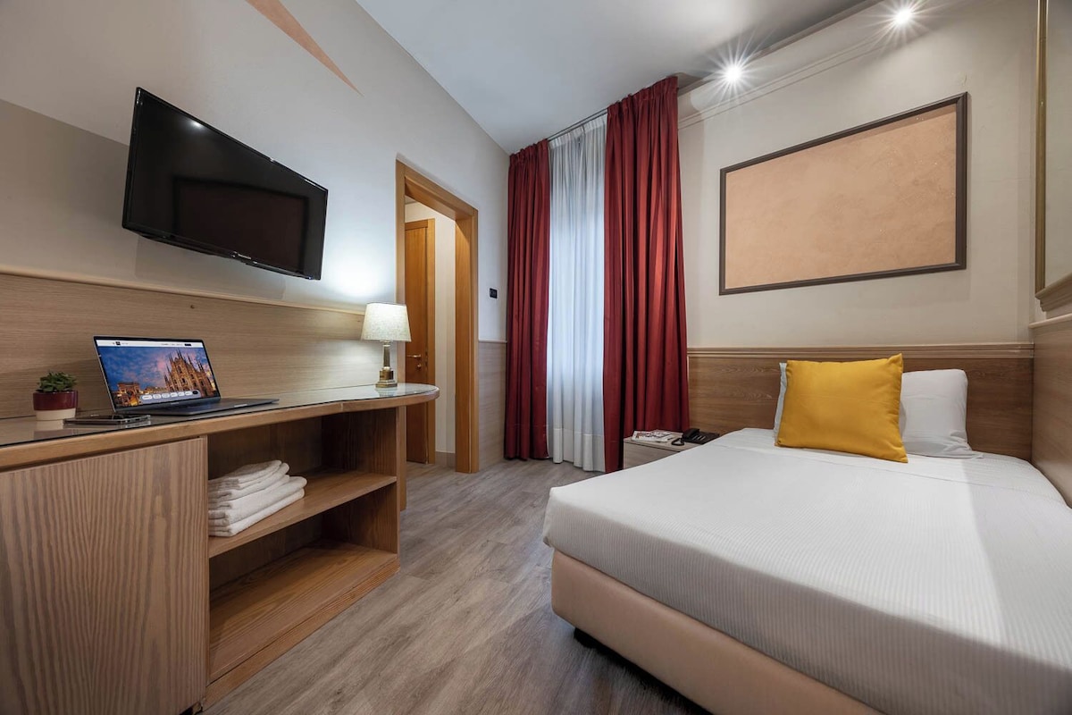 Une chambre d’hôtel avec un lit, un bureau avec un ordinateur portable, un téléviseur fixé au mur et une grande fenêtre avec des rideaux rouges.