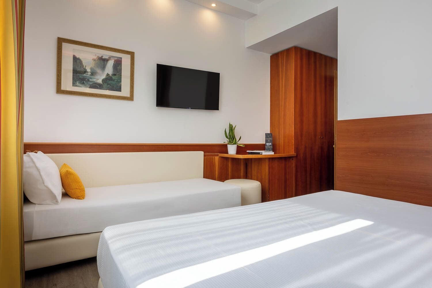 A hotel room with a bed, a sofa, a TV mounted on the wall, and a wooden wardrobe.