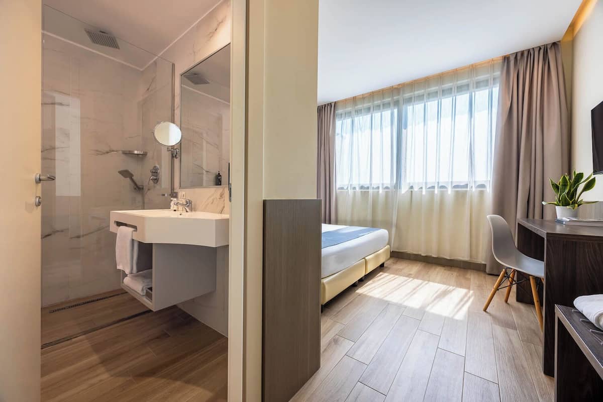 Ein Hotelzimmer mit Dusche, Waschbecken, Spiegel, Bett, Schreibtisch, Stuhl und einem Blumentopf.
