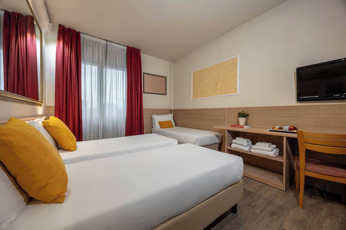 Ein Hotelzimmer mit zwei Betten, einem Schreibtisch mit Stuhl, einem Fernseher und einem Fenster mit roten Vorhängen.