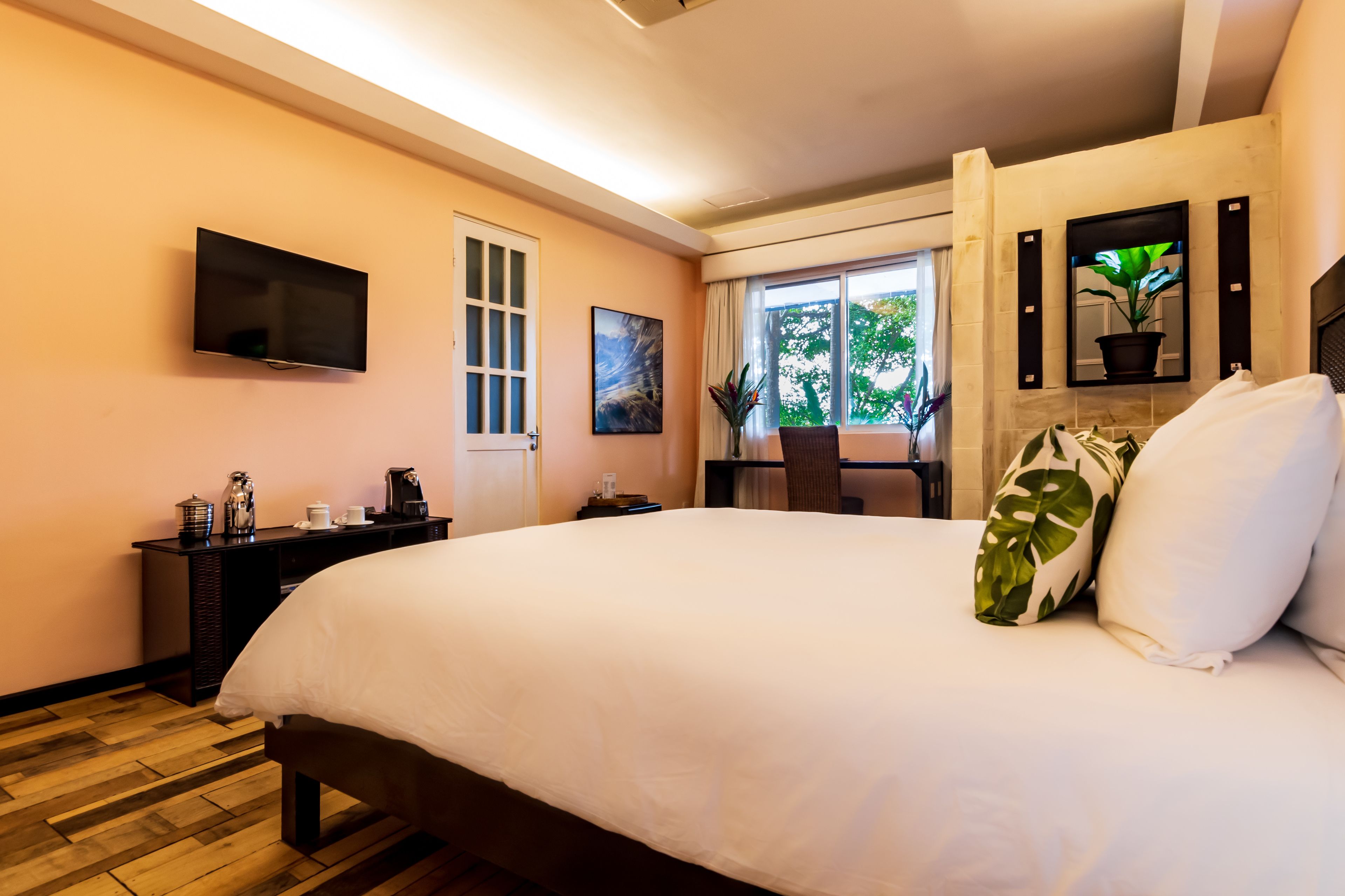 A hotel room with a large bed, a flat-screen TV mounted on the wall, a desk with a chair, and a window with a view of greenery.