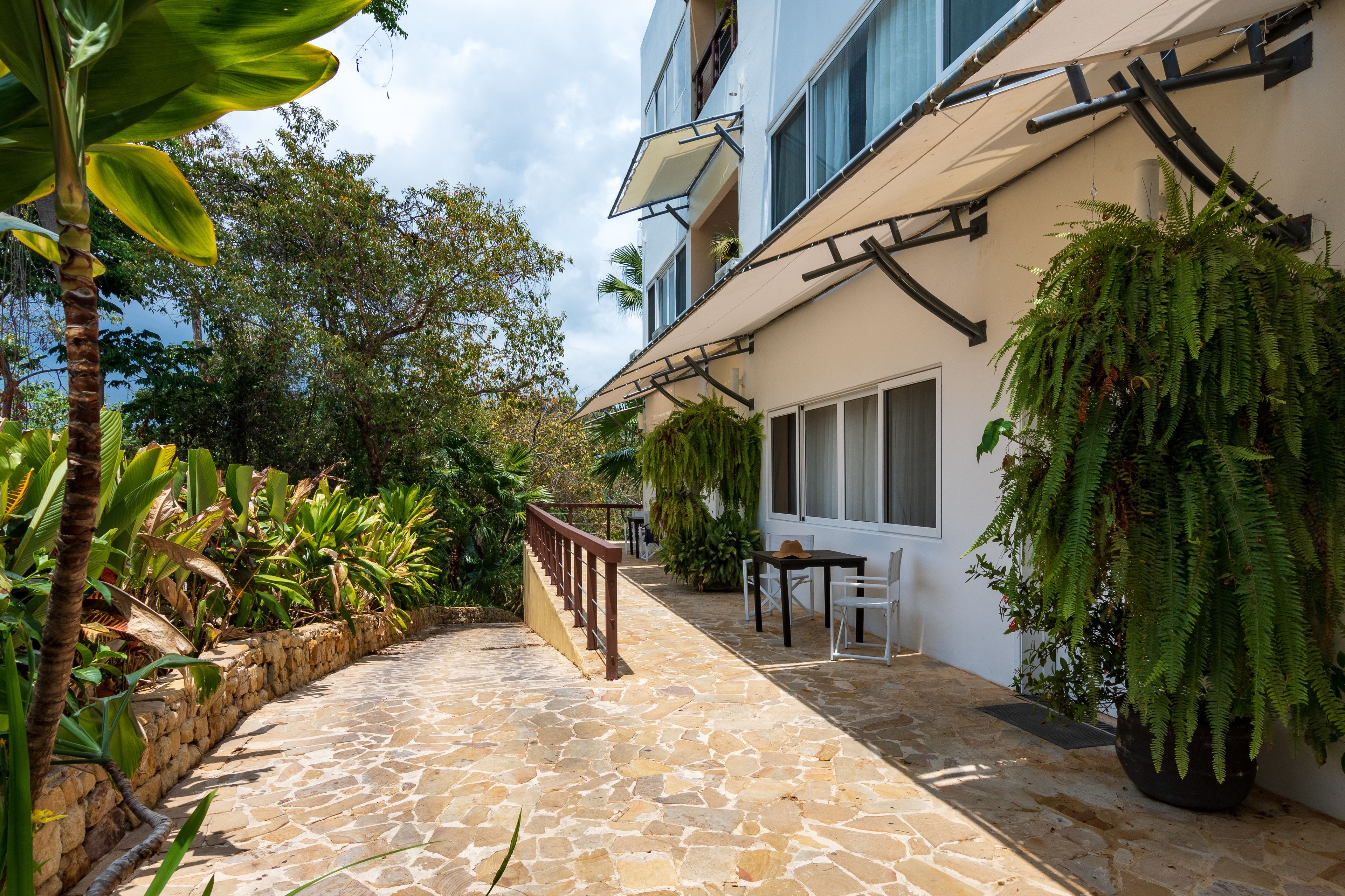 Un sentier pavé de pierres mène à un bâtiment doté d’un balcon, qui est entouré d’une végétation luxuriante et d’une plante de fougère suspendue.