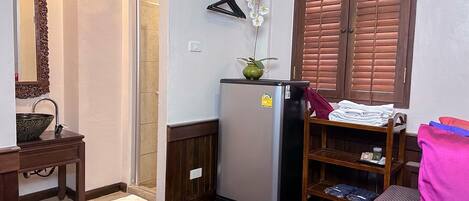 Una habitación compacta con piso de madera, un lavamanos con un cuenco decorativo, un espejo, un refrigerador, una silla de madera con un cojín rosa y una ventana con persianas de madera.