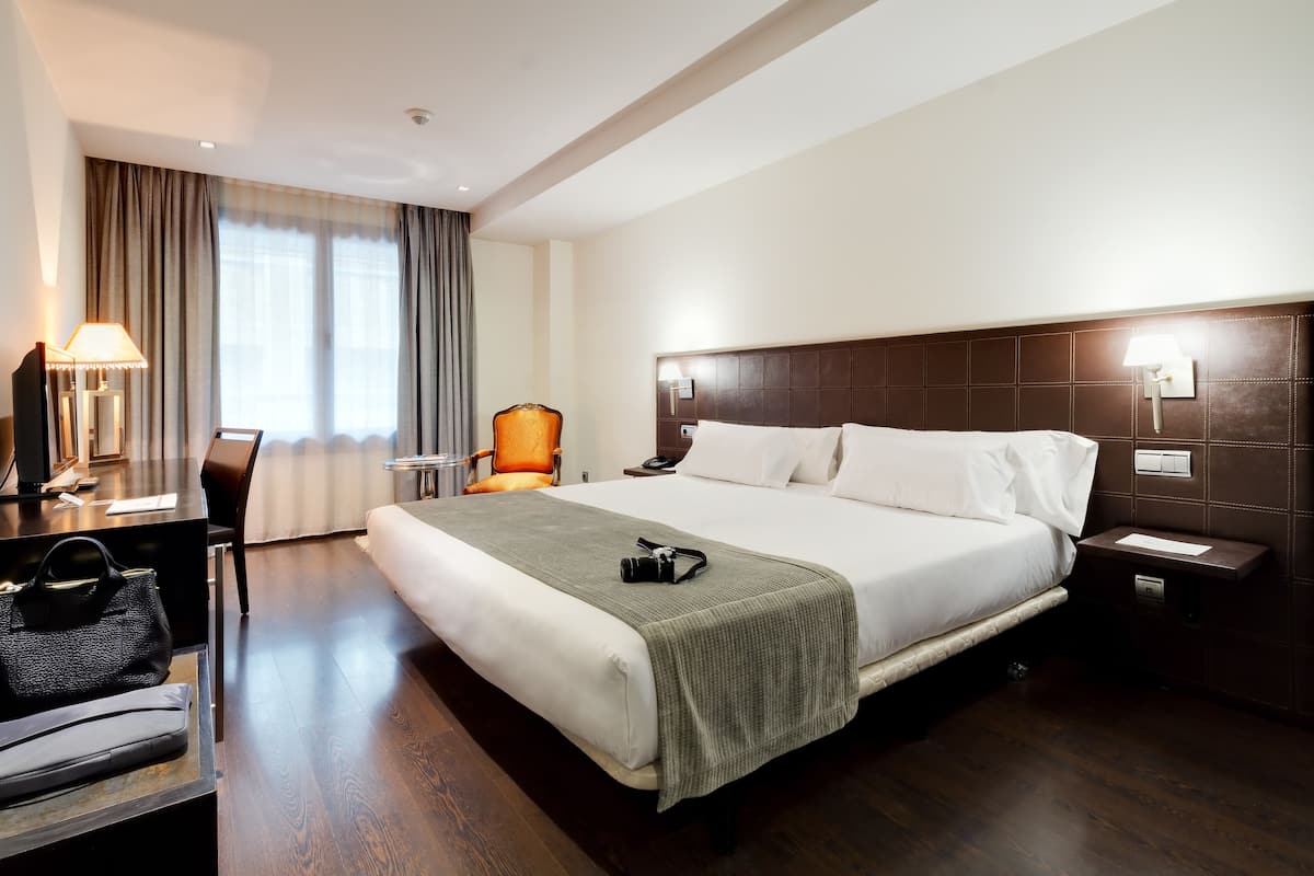Quarto de hotel com uma cama grande, uma escrivaninha com abajur, uma cadeira e uma mesa de cabeceira com telefone e luminária.