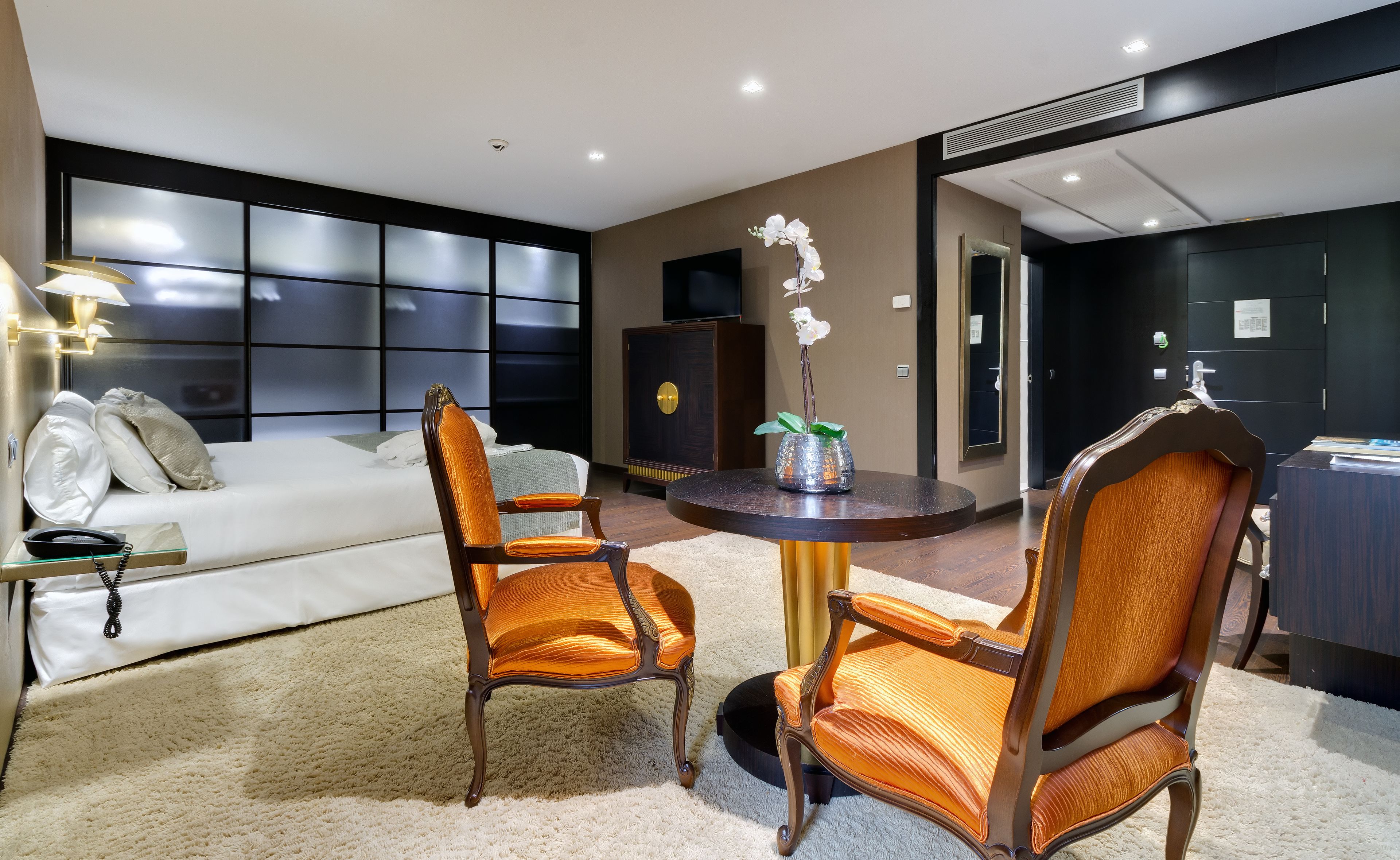 Une chambre d’hôtel moderne avec un grand lit, deux fauteuils rembourrés, une table ronde et un vase décoratif contenant une orchidée.