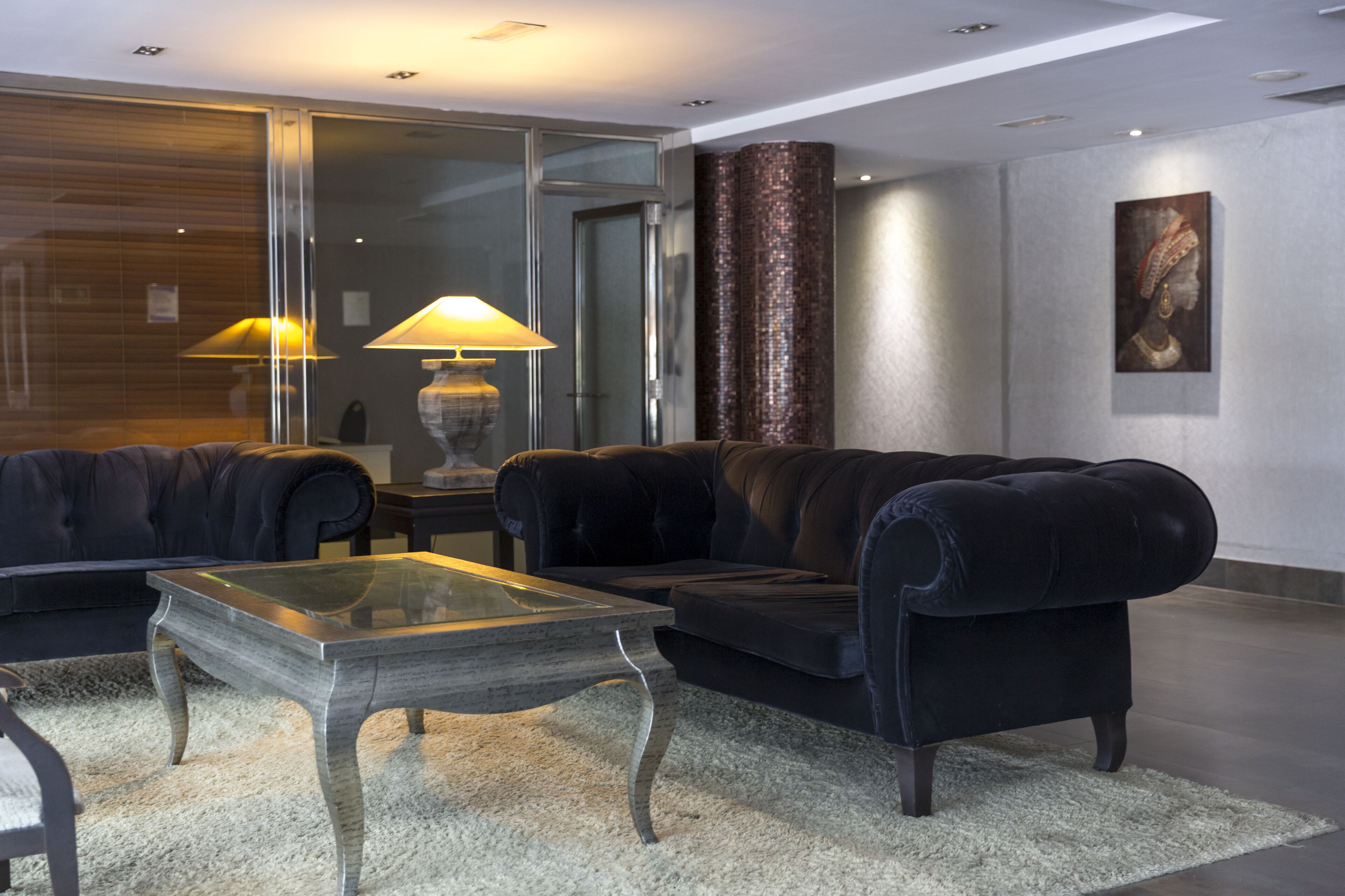A modern living room with a glass coffee table, two black sofas, a lamp, and a painting on the wall.