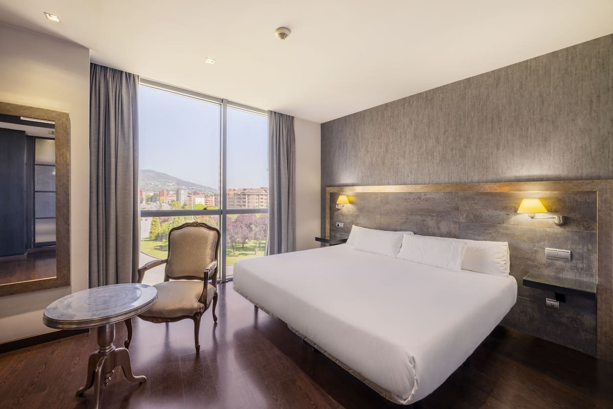 Quarto de hotel com uma cama grande, uma cadeira, uma mesinha e vista da cidade através de grandes janelas.
