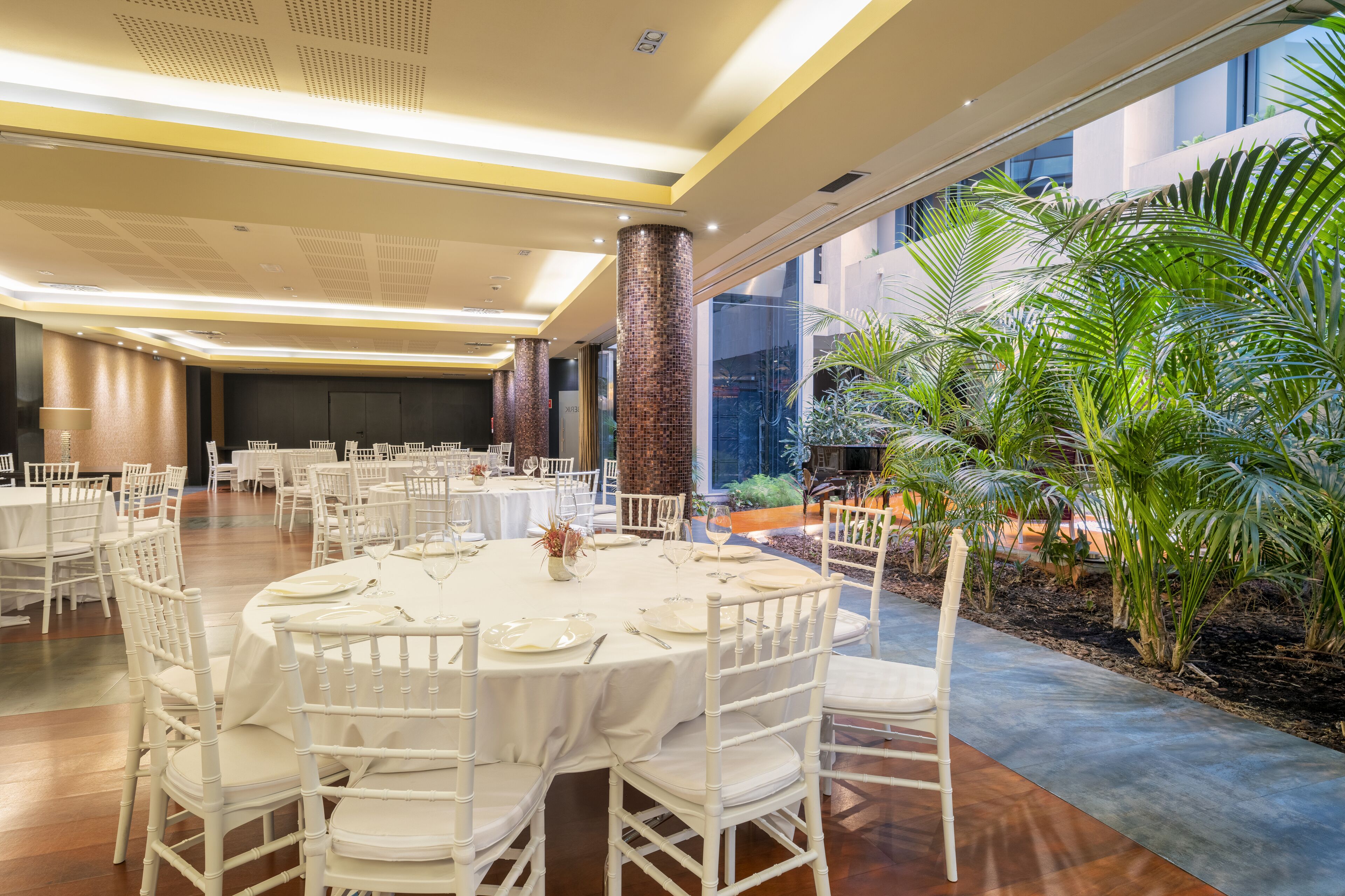 A spacious indoor dining area with round tables and white chairs, large windows, and a view of a landscaped outdoor area with palm trees.