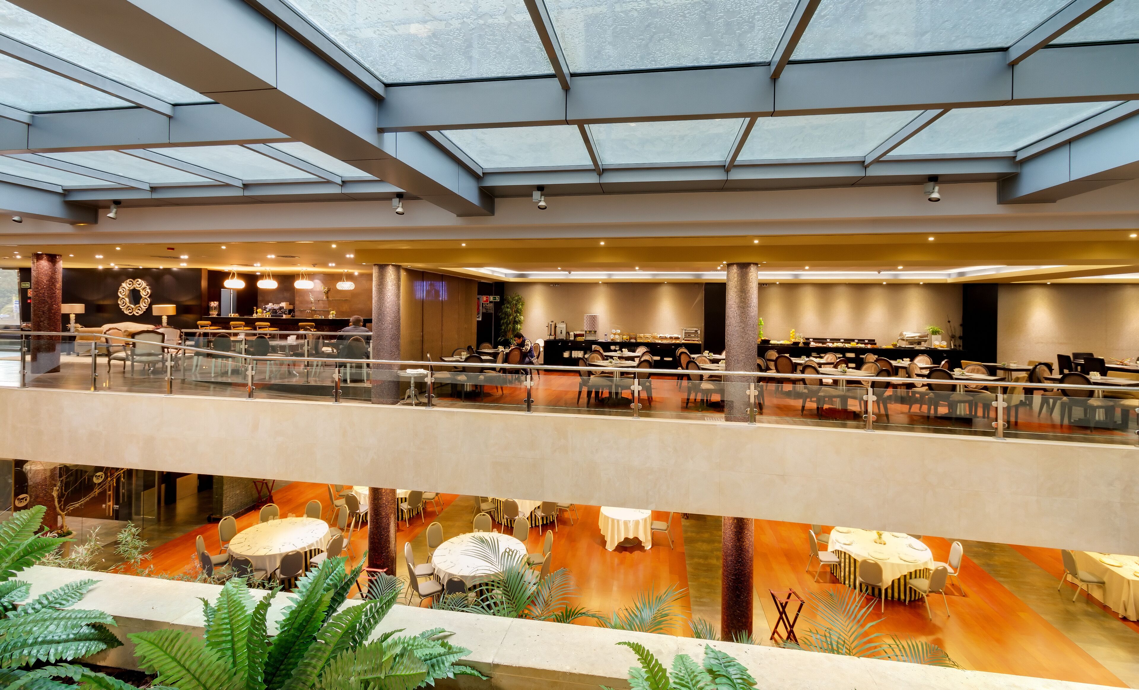 En rummelig hotellobby med flere spiseområder, et glastag og indendørs planter.