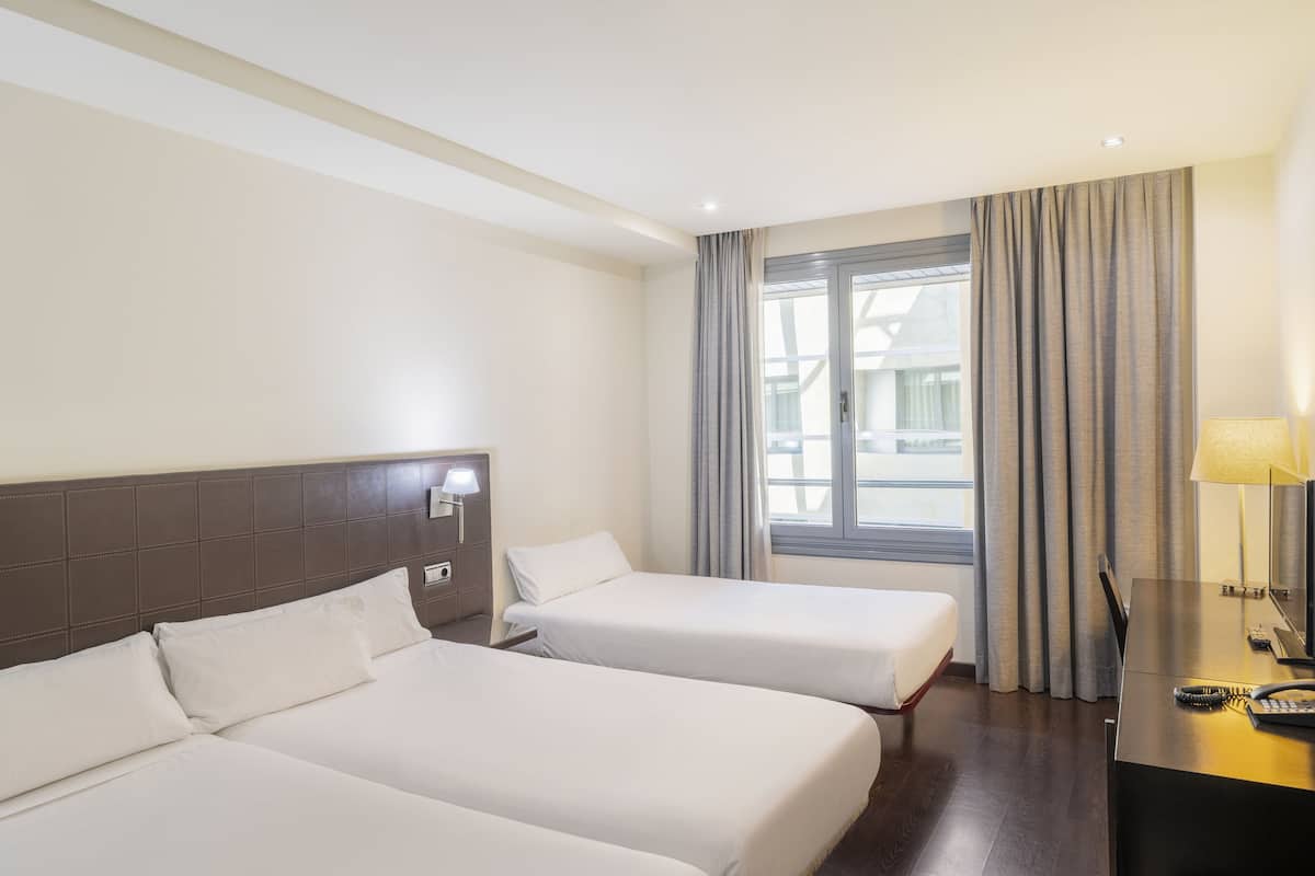 Quarto de hotel com duas camas, uma escrivaninha com abajur e uma janela com cortinas.