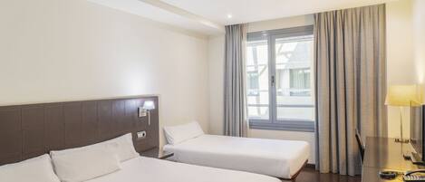 Una habitación de hotel con dos camas, un escritorio con lámpara y una ventana con cortinas.