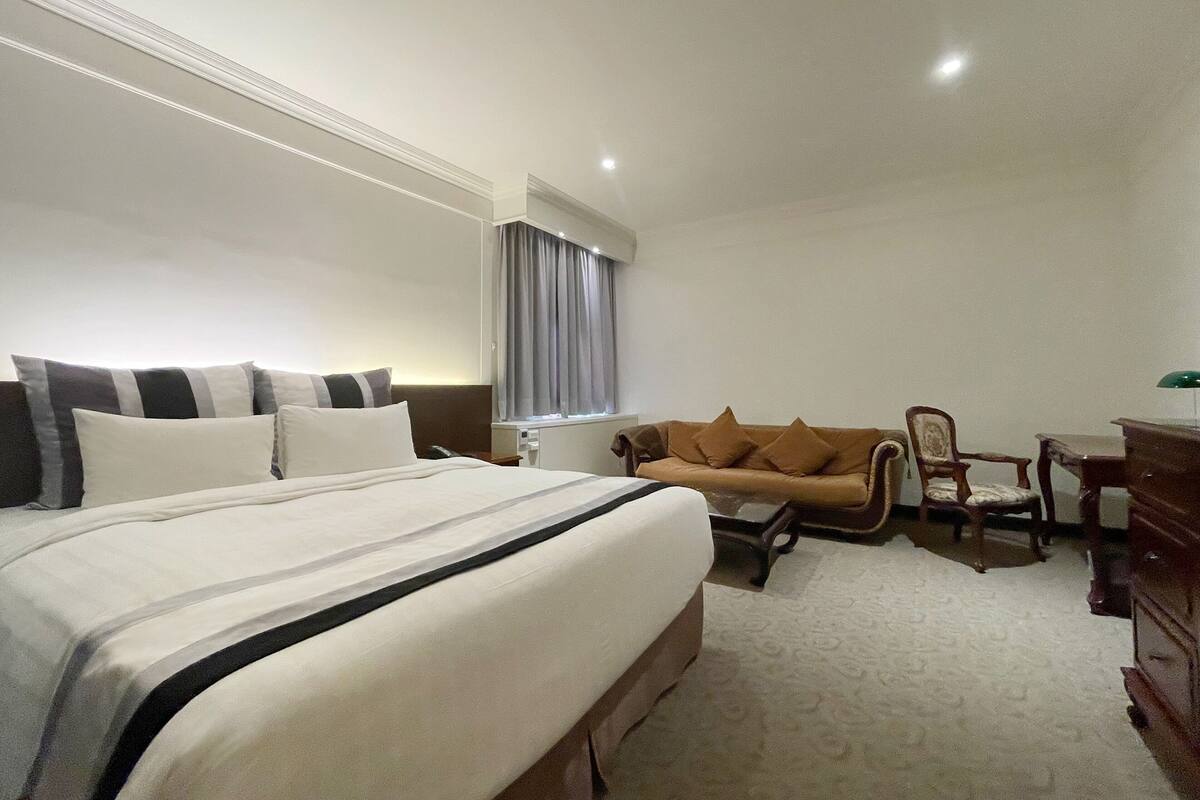 Camera d'albergo con un letto grande, un divano marrone, una scrivania con una lampada e una finestra con tende.