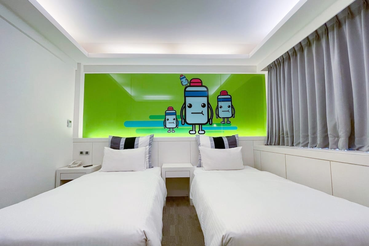 Una camera d'albergo con due letti, una parete verde decorata con personaggi dei cartoni animati, una finestra con tende e un comodino con un telefono.