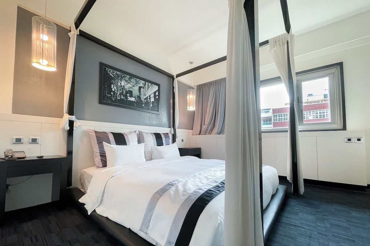 Camera d'albergo moderna con un letto grande, una testiera con un dipinto di skyline urbano, un comodino con un telefono e vista sugli edifici dalla finestra.