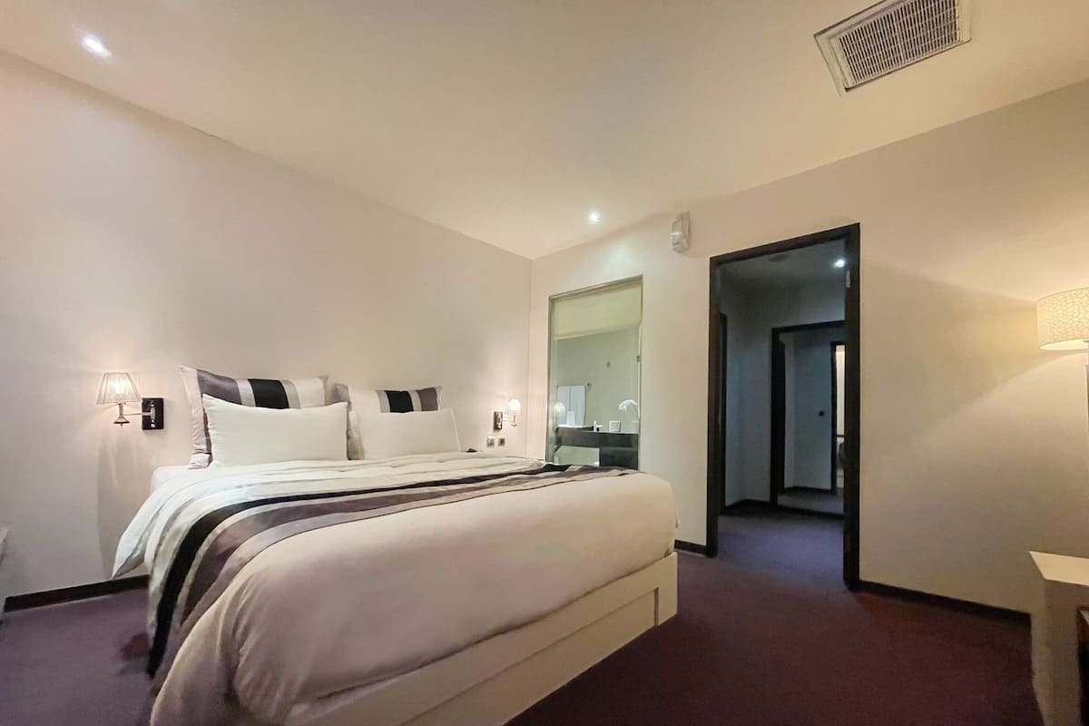 Una camera d'albergo con un letto grande, lampade ai comodini, uno specchio e una porta che dà su un'altra stanza.