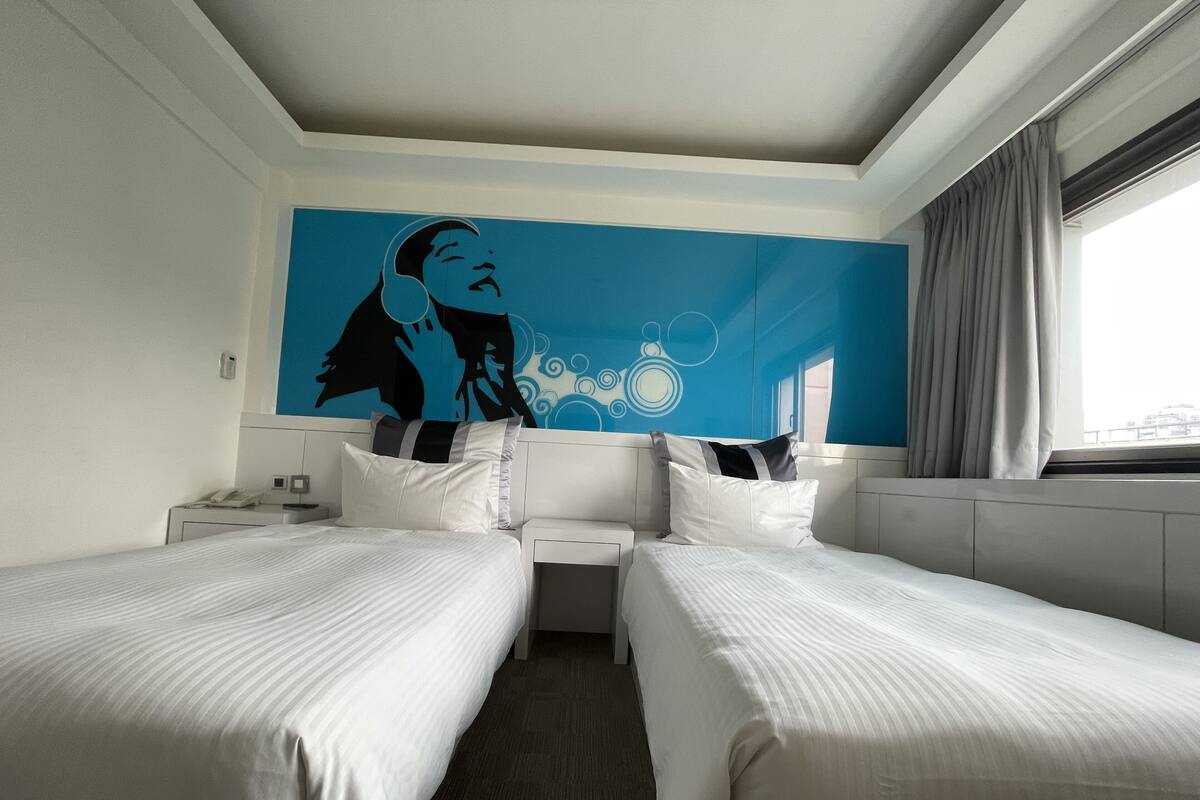 Camera d'albergo con due letti, un'ampia finestra con tende, un murales a parete e un comodino con il telefono.